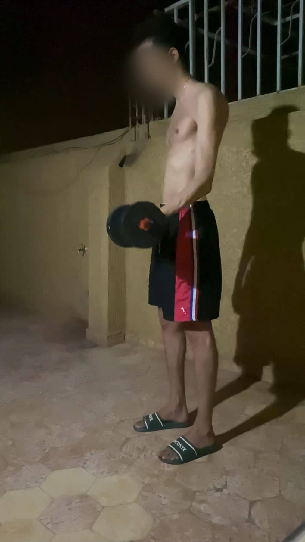 Mehdi alone on a roof at night, shirtless and very thin, holding one small dumbbell