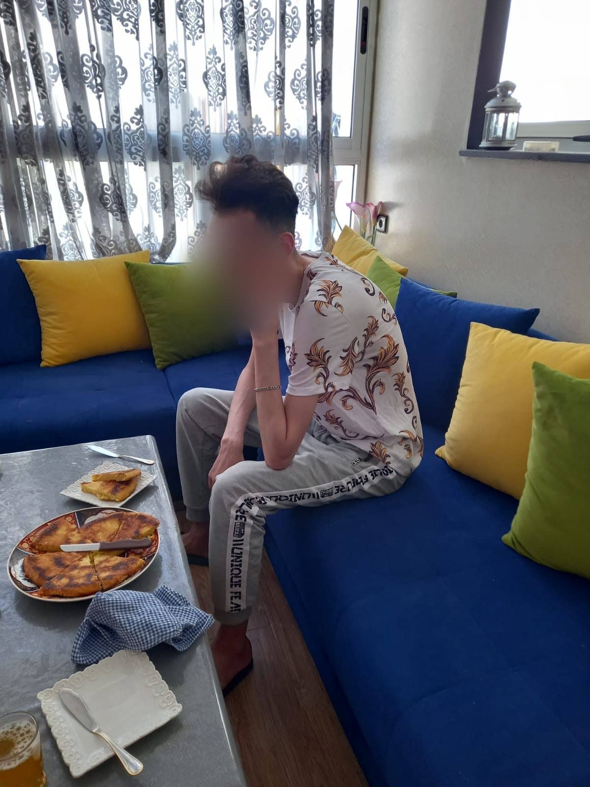 Mehdi sitting alone on a blue sofa, a plate of food left on the table